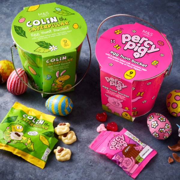 Two M&S colouful easter egg hunt buckets; one is colin the caterpillar and the other is percy pig. There are other sweets and eggs scattered around them,